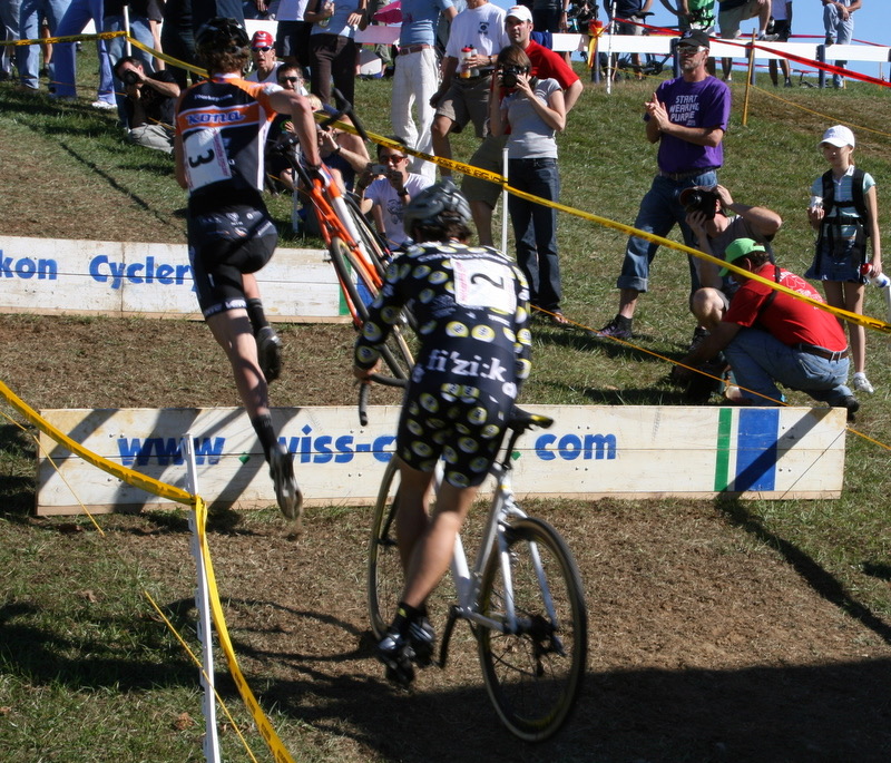 Cyclocross racers carrying bikes up a grassy slope past course tape